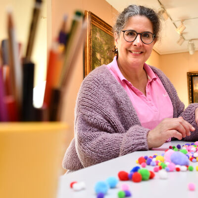 Museumspädagogin Esther Dörrhöfer sitzt an einem Basteltisch in der Dauerausstellung des Ostholstein-Museums. Während sie bunte Bastelbommeln aus einem Glas auf dem Tisch verteilt, ist im Vordergrund ein Stiftebecher mit verschiedenen Stiften in künstlerischer Unschärfe zu sehen. Im Hintergrund heben sich die historischen Exponate der Ausstellung ab.